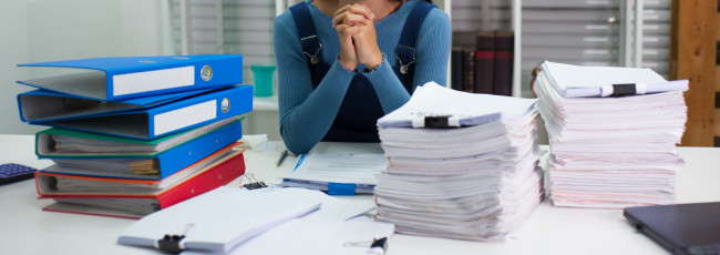 A woman clasps her hands at the center of two huge piles of paperwork.